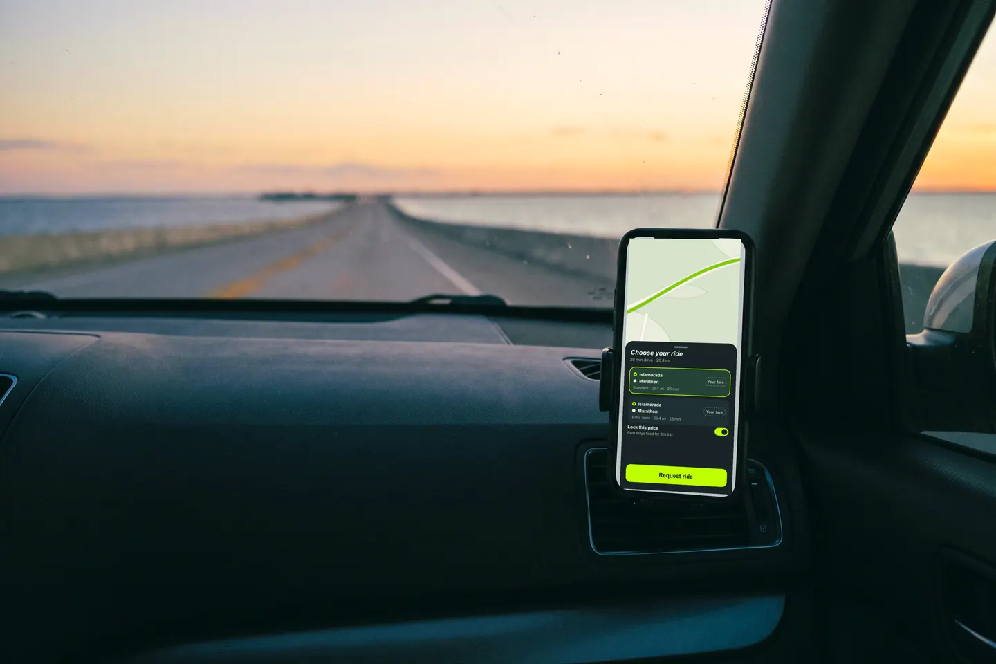 A phone in a dashboard mount showing the Outride rider app, the causeway ahead at dusk