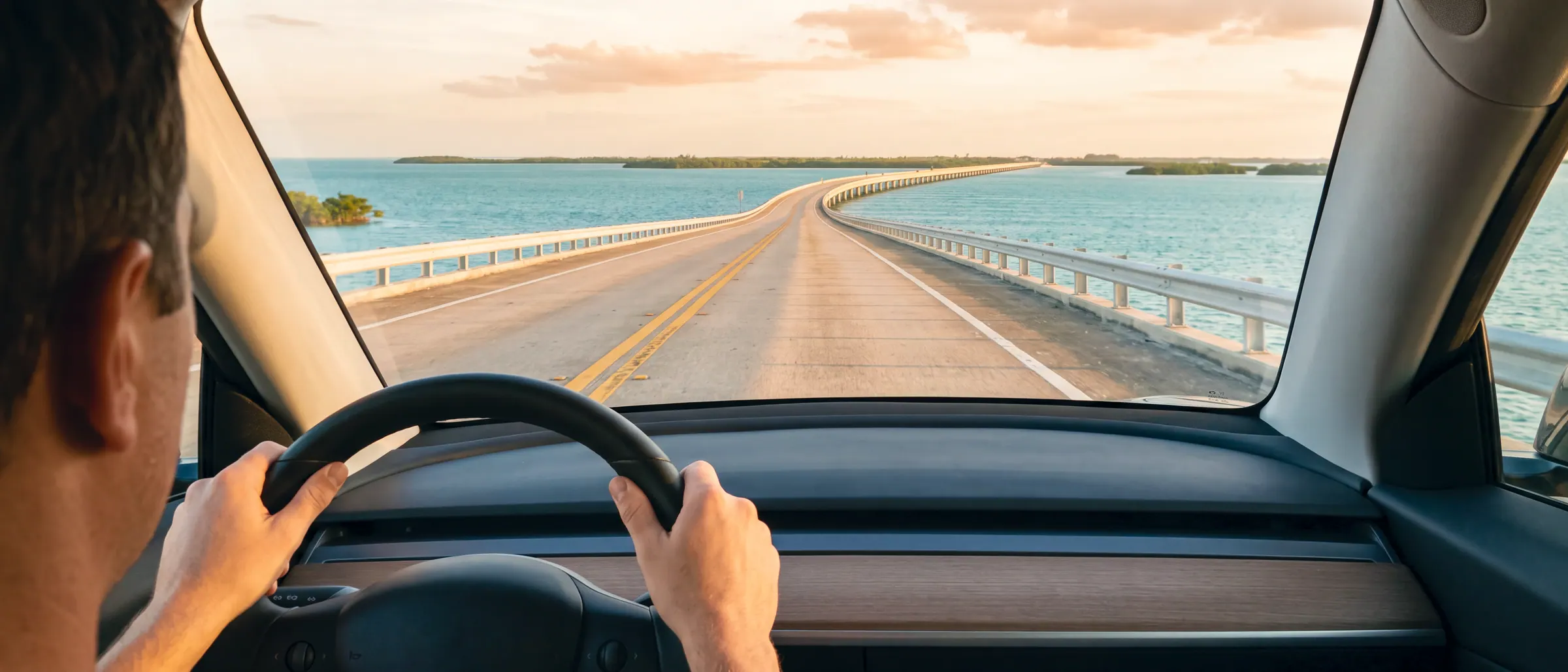 Hands on the wheel of a car crossing a causeway over open water at dusk